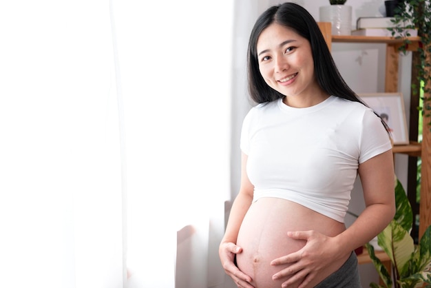 Jovem mulher grávida bonita asiática segura as mãos na barriga em pé perto da janela em casa Preparação para maternidade de gravidez e conceito de expectativa