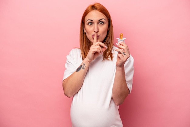 Jovem mulher grávida caucasiana segurando chupeta isolada em fundo rosa mantendo um segredo ou pedindo silêncio