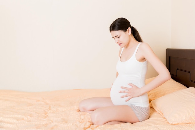 Jovem mulher grávida com dor sentada na cama no quarto se preparando para dar à luz