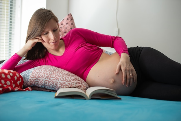 Jovem mulher grávida deitada em uma cama lendo um livro.