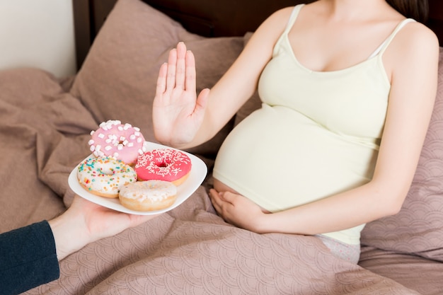 Jovem mulher grávida deitada na cama, recusando rosquinhas