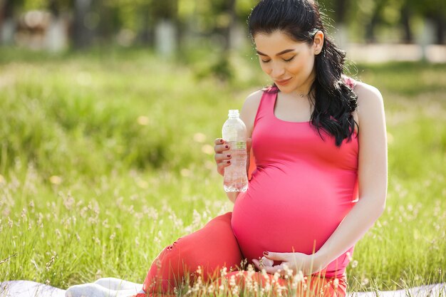 Jovem mulher grávida fazendo exercícios de fitness ao ar livre e bebendo água mineral limpa.