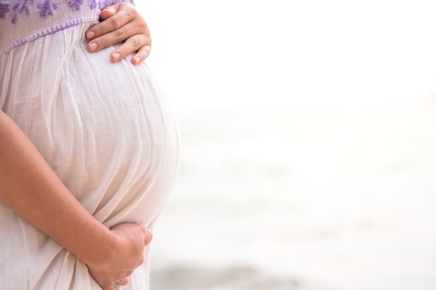 Jovem mulher grávida no vestido de mãos dadas na barriga com fundo do mar turva