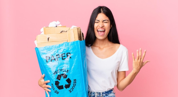Jovem mulher hispânica bonita gritando agressivamente com muita raiva e segurando um saco de papel para reciclar
