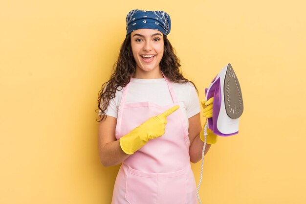 Jovem mulher hispânica bonita olhando animada e surpresa apontando para o conceito de ferro e roupas de lado