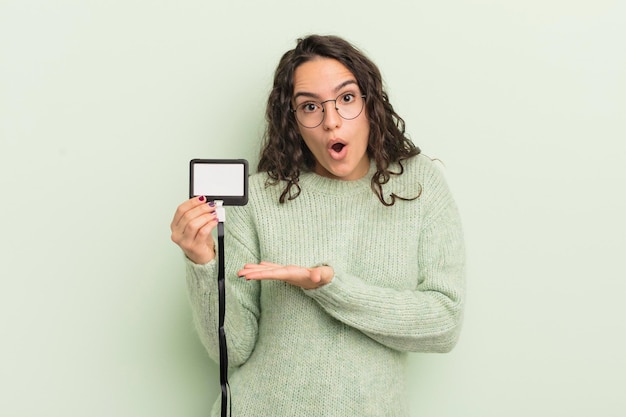 Jovem mulher hispânica bonita parecendo surpresa e chocada com o queixo caído segurando uma identificação de cartão de credenciamento de objeto