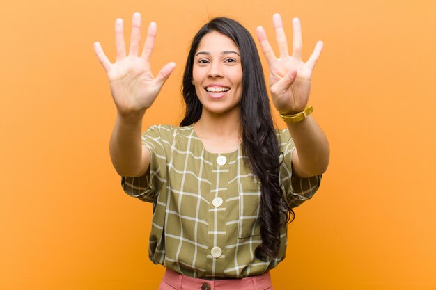 Foto jovem mulher hispânica bonita sorrindo e olhando amigável, mostrando o número nove ou nono com a mão para a frente, contando para baixo contra a parede marrom