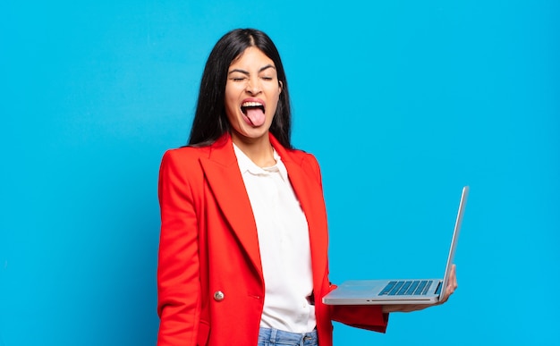 Jovem mulher hispânica com atitude alegre, despreocupada, rebelde, brincando e mostrando a língua, se divertindo. conceito de laptop