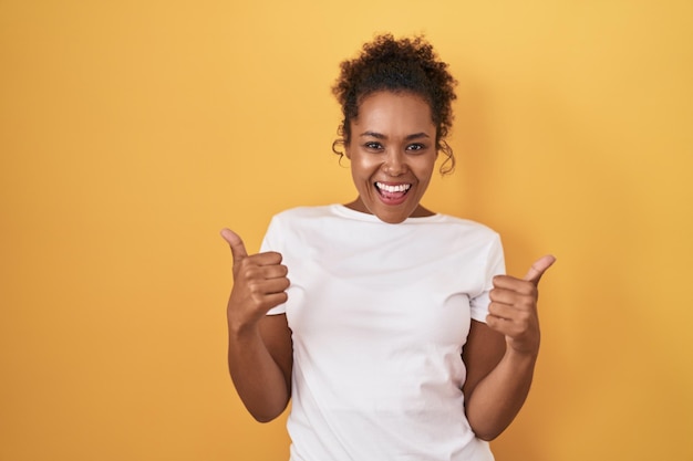 Jovem mulher hispânica com cabelo encaracolado em pé sobre sinal de sucesso de fundo amarelo fazendo gesto positivo com a mão, polegares para cima sorrindo e feliz. expressão alegre e gesto vencedor.