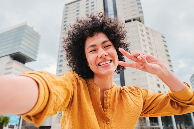 Jovem mulher hispânica com camisa amarela de cabelo encaracolado e atitude alegre sorrindo e se divertindo tirando uma foto de selfie fazendo o sinal de paz com os dedos na rua da cidade ao ar livre Conceito de estilo de vida