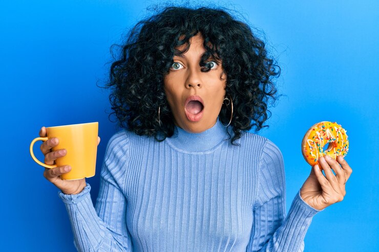 Foto jovem mulher hispânica comendo rosquinha e bebendo café com medo e chocada com surpresa e expressão espantada, medo e rosto animado.