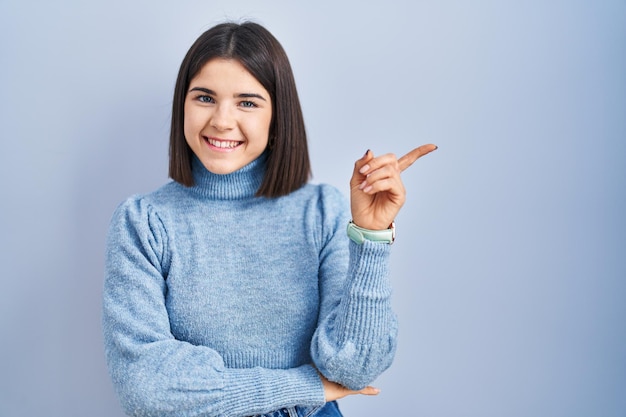 Jovem mulher hispânica em pé sobre fundo azul com um grande sorriso no rosto apontando com o dedo da mão para o lado olhando para a câmera