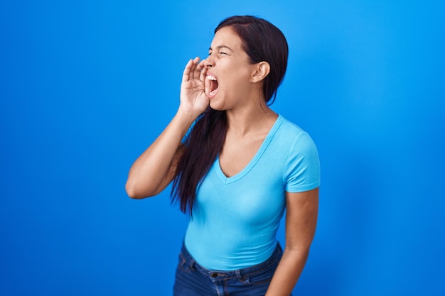 Jovem mulher hispânica em pé sobre fundo azul gritando e gritando alto a lado com a mão no conceito de comunicação da boca
