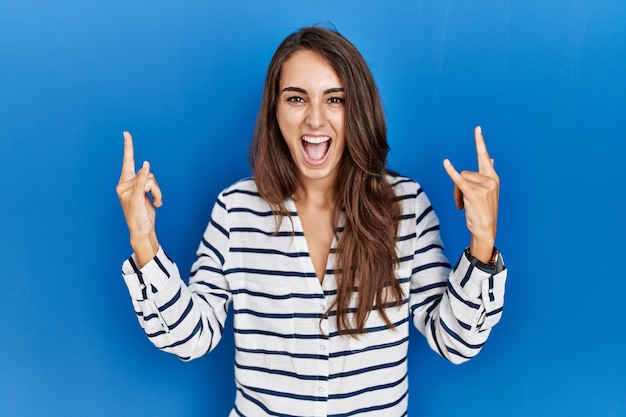 Jovem mulher hispânica em pé sobre fundo azul isolado gritando com expressão maluca fazendo símbolo de rock com as mãos para cima. estrela da música. conceito de música pesada.