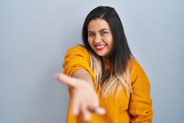 Jovem mulher hispânica em pé sobre fundo isolado sorrindo alegre oferecendo mão de palma dando assistência e aceitação