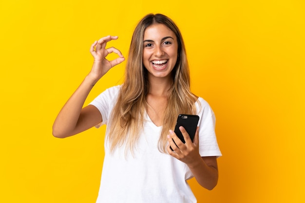 Jovem mulher hispânica em uma parede amarela isolada usando telefone celular e fazendo sinal de ok