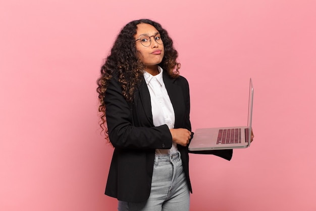 Jovem mulher hispânica encolhendo os ombros, sentindo-se confusa e incerta, duvidando com os braços cruzados e olhar perplexo. conceito de laptop