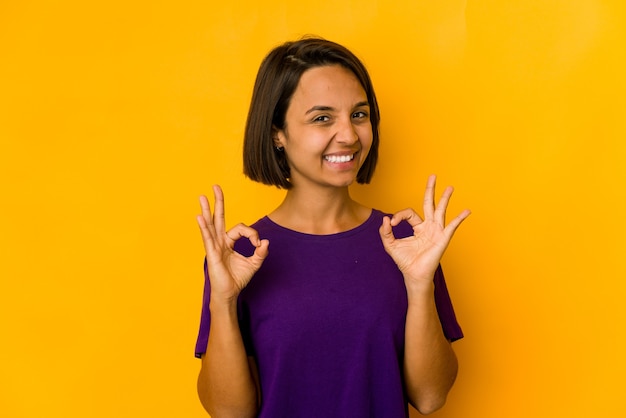 Jovem mulher hispânica isolada em amarelo alegre e confiante, mostrando um gesto de ok.