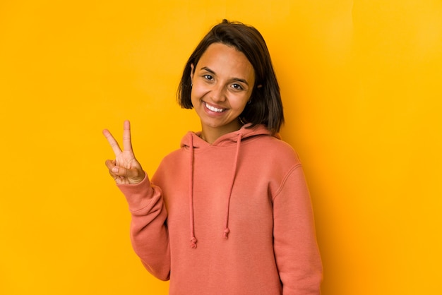 Jovem mulher hispânica isolada em amarelo mostrando sinal de vitória e sorrindo amplamente.