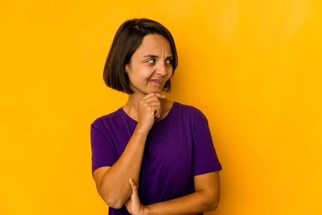 Foto jovem mulher hispânica isolada em amarelo, olhando de soslaio com expressão duvidosa e cética.