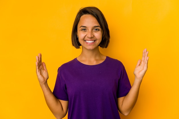 Jovem mulher hispânica isolada em amarelo segurando algo pequeno com os indicadores, sorrindo e confiante.