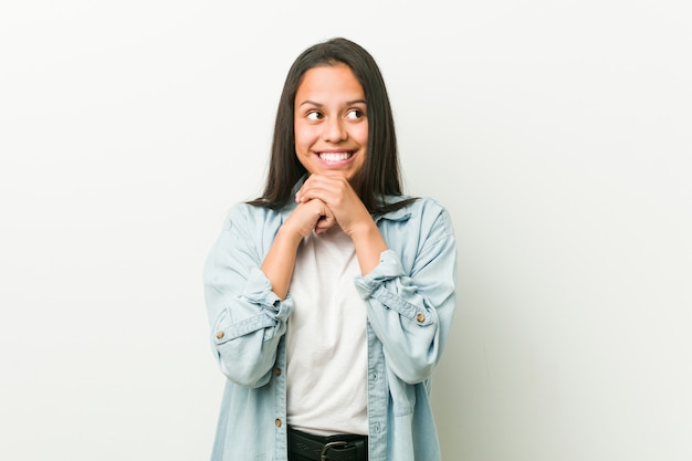 Jovem mulher hispânica mantém as mãos sob o queixo, está olhando alegremente de lado.