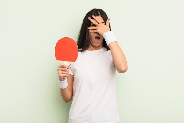 Jovem mulher hispânica olhando chocada, assustada ou aterrorizada, cobrindo o rosto com o conceito de ping pong de mão