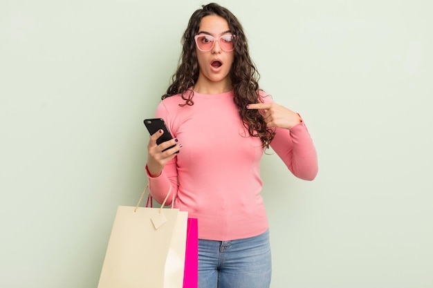 Jovem mulher hispânica olhando chocada e surpresa com a boca aberta apontando para o conceito de sacolas de compras