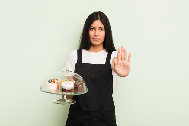 Foto jovem mulher hispânica olhando séria mostrando a palma da mão aberta fazendo gesto de parada chef com conceito de bolos