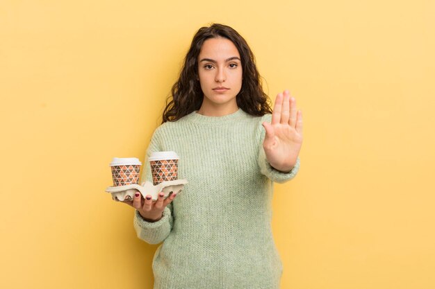 Jovem mulher hispânica olhando séria mostrando a palma da mão aberta, fazendo o gesto de parada. take away conceito de café