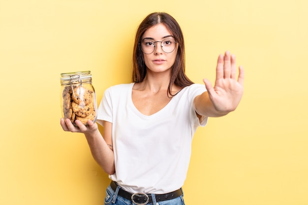 Jovem mulher hispânica olhando sério, mostrando a palma da mão aberta, fazendo o gesto de parada. conceito de garrafa de biscoitos