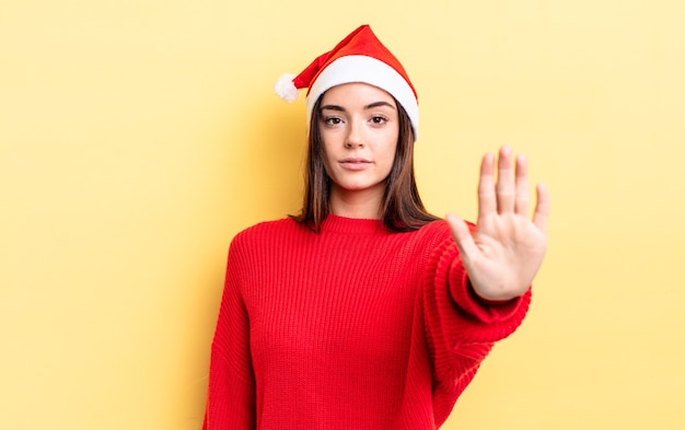 Jovem mulher hispânica olhando sério, mostrando a palma da mão aberta, fazendo o gesto de parada. Natal e conceito de ano novo