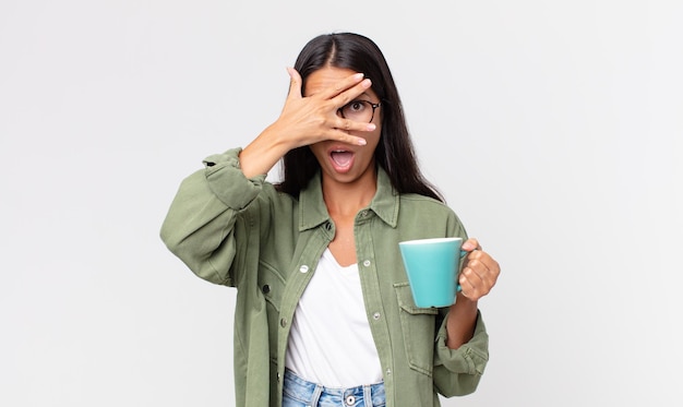 Jovem mulher hispânica parecendo chocada, assustada ou apavorada, cobrindo o rosto com a mão e segurando uma caneca de café