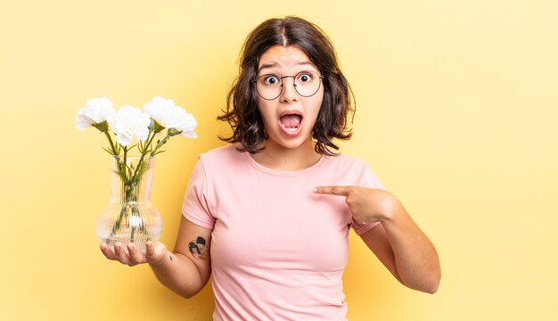 Jovem mulher hispânica parecendo chocada e surpresa com a boca aberta, apontando para si mesma. conceito de vaso de flores