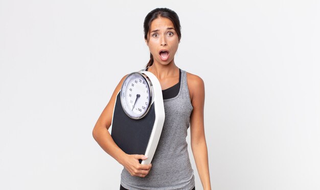 Jovem mulher hispânica parecendo muito chocada ou surpresa e segurando uma balança. conceito de dieta