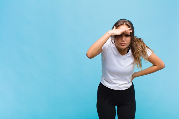 Jovem mulher hispânica parecendo perplexa e atônita, com a mão na testa olhando para longe, observando ou procurando