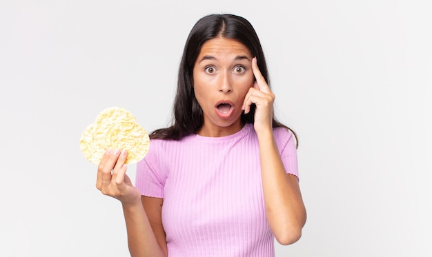 Jovem mulher hispânica parecendo surpresa, percebendo um novo pensamento, ideia ou conceito e segurando um biscoito de arroz. conceito de dieta