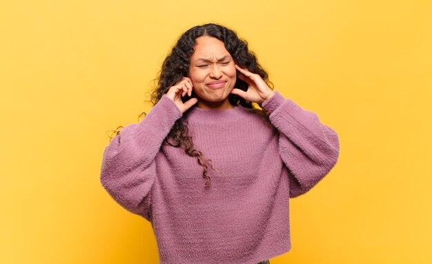 Jovem mulher hispânica parecendo zangada, estressada e irritada, cobrindo ambos os ouvidos para um barulho, som ou música alta ensurdecedores