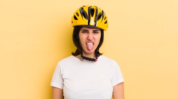 Jovem mulher hispânica se sentindo enojada e irritada e com a língua para fora do conceito de capacete de bicicleta