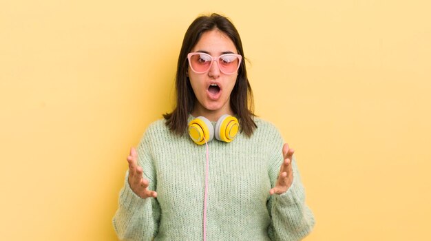 Jovem mulher hispânica se sentindo extremamente chocado e surpreso com o conceito de fones de ouvido e óculos de sol