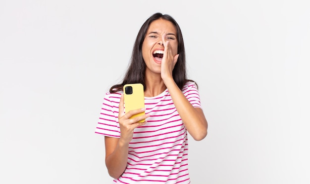 Jovem mulher hispânica se sentindo feliz, dando um grande grito com as mãos perto da boca e segurando um smartphone