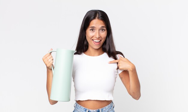 Jovem mulher hispânica se sentindo feliz e apontando para si mesma com uma garrafa térmica de café animada e segurando