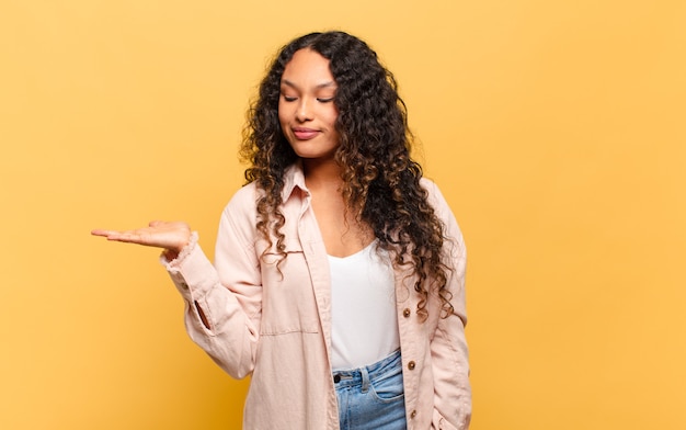 Jovem mulher hispânica se sentindo feliz e sorrindo casualmente, olhando para um objeto ou conceito seguro ao lado