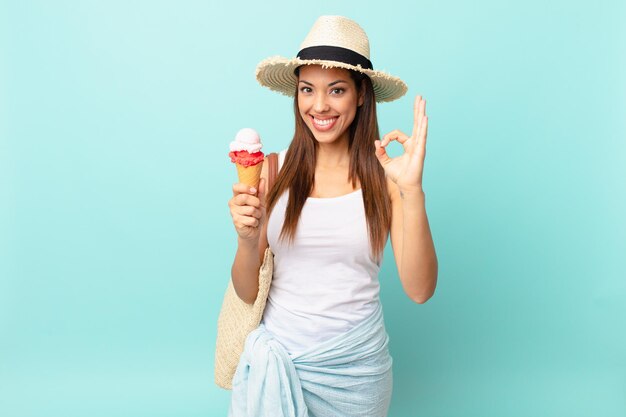 Jovem mulher hispânica se sentindo feliz, mostrando aprovação com um gesto ok e segurando um sorvete. conceito de sumer