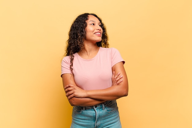 Jovem mulher hispânica se sentindo feliz, orgulhosa e esperançosa, imaginando ou pensando, olhando para cima para copiar o espaço com os braços cruzados