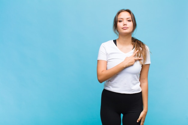 Jovem mulher hispânica se sentindo feliz, positiva e bem-sucedida, com a mão fazendo formato de V no peito, mostrando paz