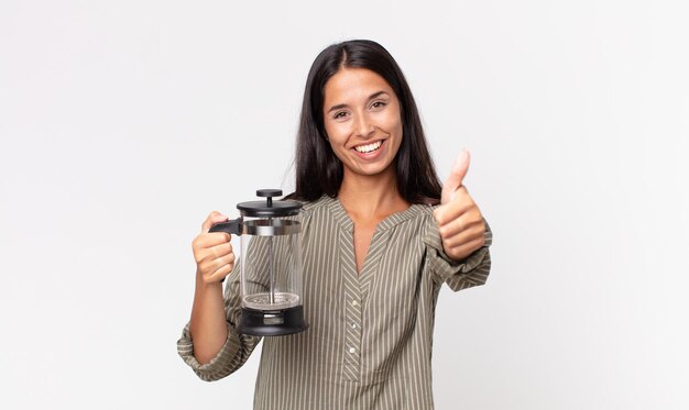 Jovem mulher hispânica se sentindo orgulhosa, sorrindo positivamente com os polegares para cima e segurando uma cafeteira manual