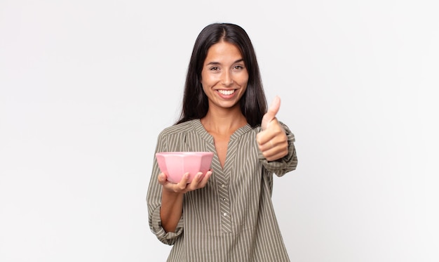 Jovem mulher hispânica se sentindo orgulhosa sorrindo positivamente com os polegares para cima e segurando uma tigela ou pote vazio