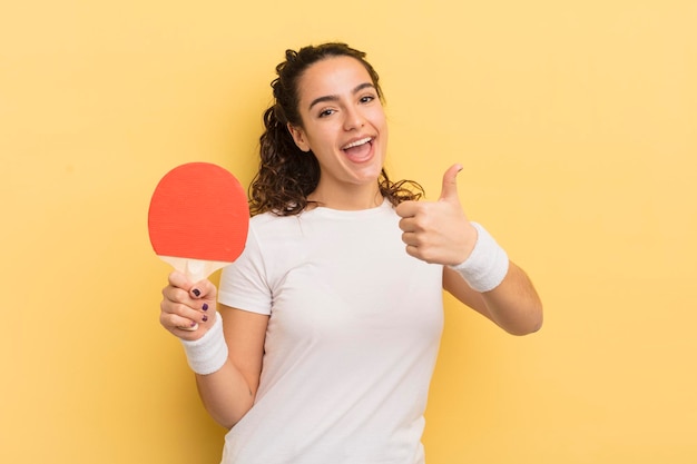 Jovem mulher hispânica se sentindo orgulhosa sorrindo positivamente com polegares para cima conceito de pingue-pongue