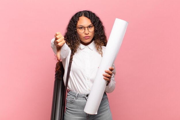 Jovem mulher hispânica se sentindo zangada, irritada, decepcionada ou descontente, mostrando os polegares para baixo com um olhar sério. conceito de arquiteto